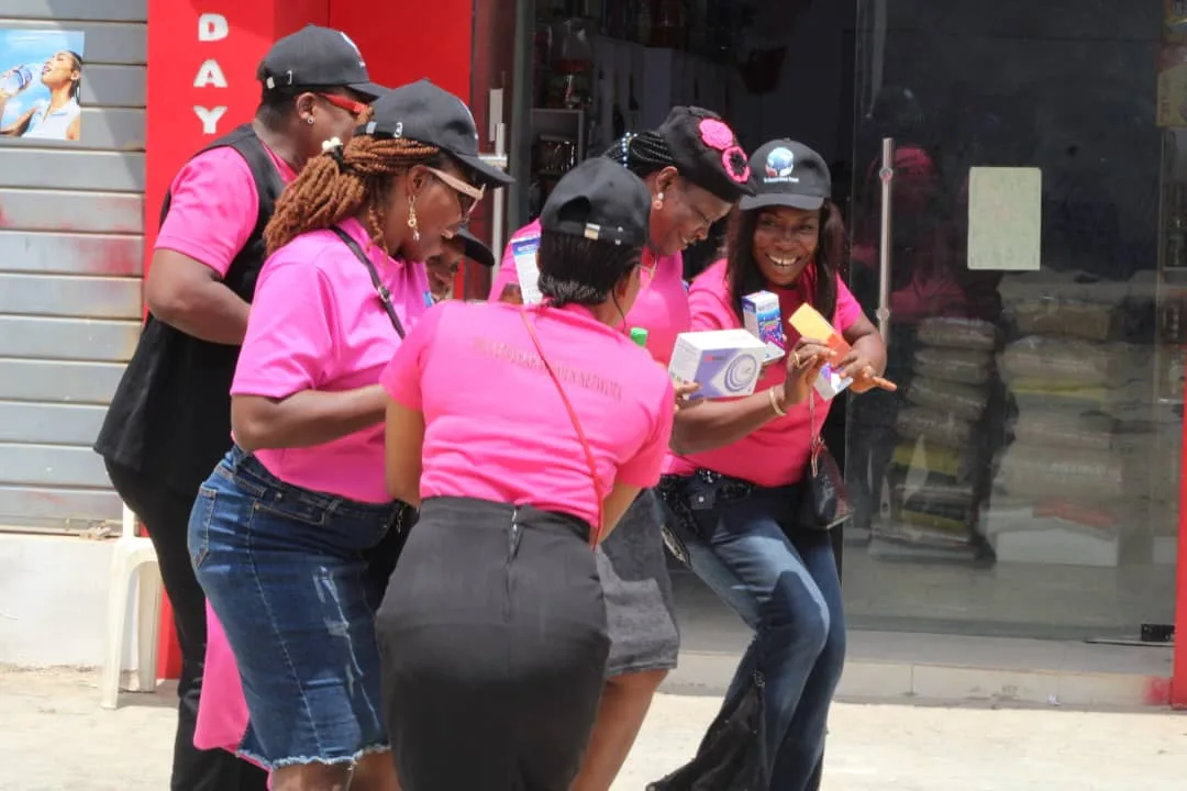Members in pink shirts laughing together on a street outside a shop, carrying boxes of medicine