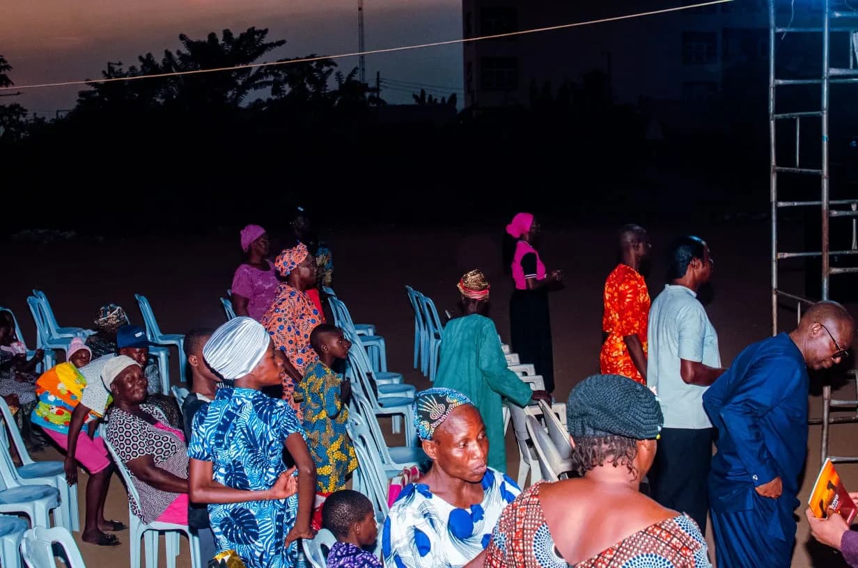Worshippers standing among rows of white plastic chairs on the open crusade ground at dusk