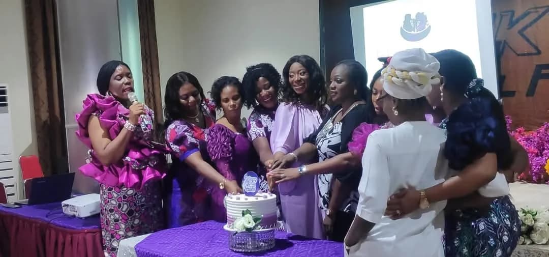 Nine women in purple gathered around a decorated cake, cutting it together