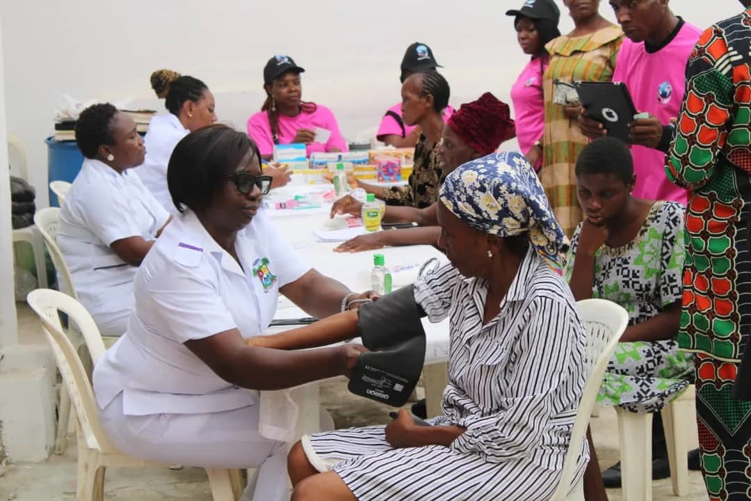 A nurse in white taking a woman’s blood pressure with a cuff, at a table of medicines outdoors