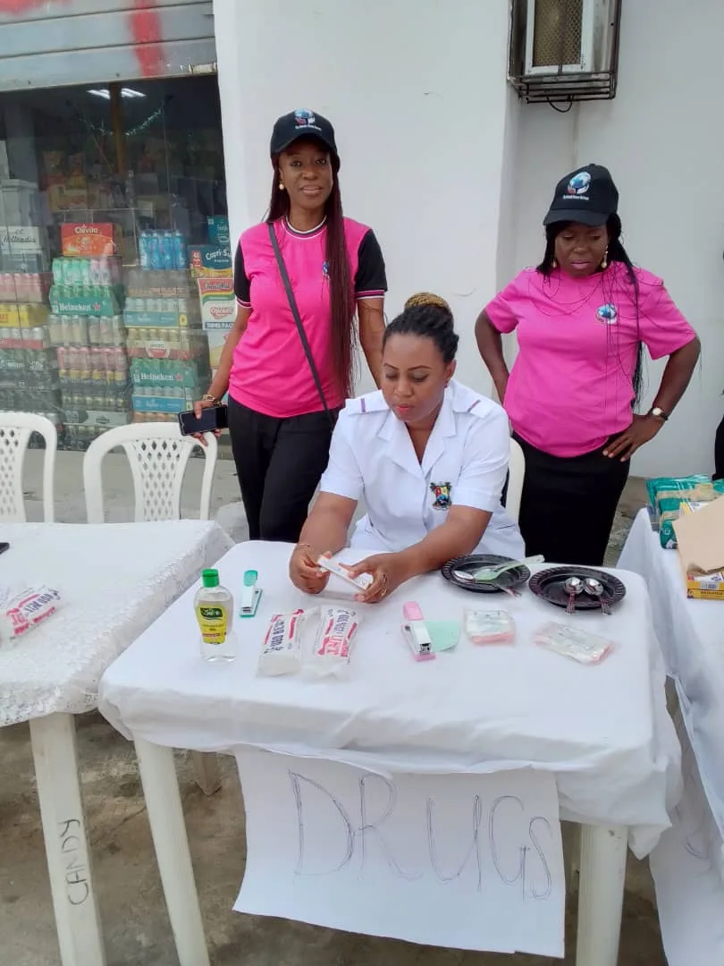 A nurse sorting medicines at a table hand-labelled “Drugs”, with two ministry members standing by