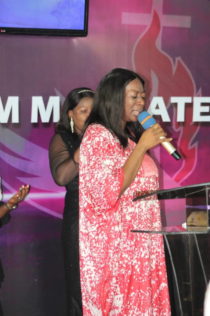 A minister in a coral gown leading prayer with her eyes closed, another standing behind