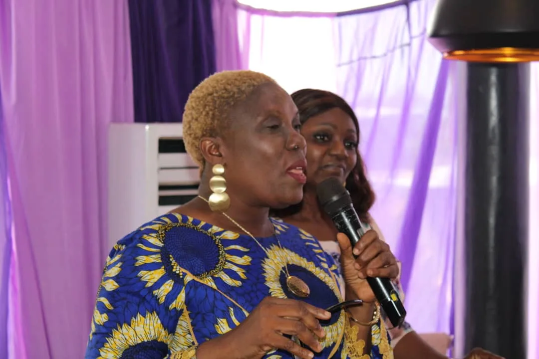 A woman in blue and gold Ankara ministering with a microphone, another standing behind her
